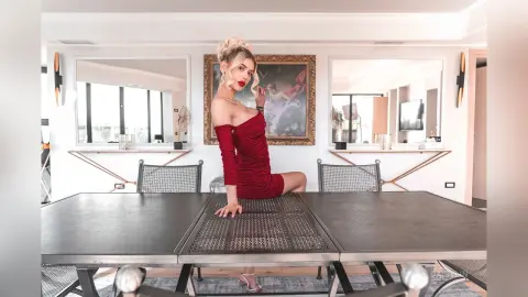 DaisyLin wears a red off-the-shoulder dress while sitting on a metal mesh table in a bright room.