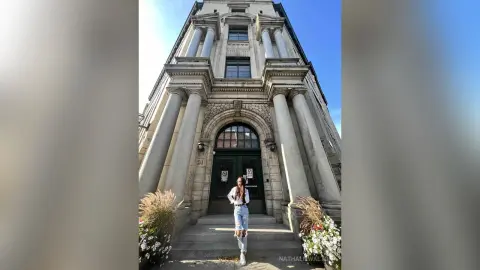 Fashionable woman in light blue jeans and white top standing before a grand neoclassical stone building.
