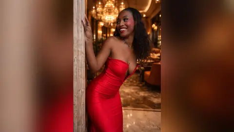 LiaTyler smiles while wearing a tight, strapless red dress, posing against a marble pillar in a dimly lit, warm room.