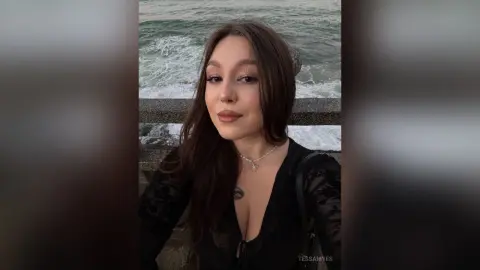Young woman in a black lace top smiling by the ocean waves at sunset.