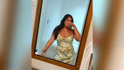 Young woman in a floral print slip dress sipping a drink in a bright bathroom mirror selfie.