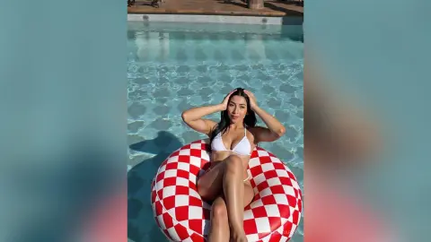Smiling woman in a white bikini relaxing on a red and white checkered float in a sunny swimming pool.
