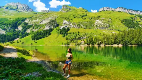 Woman in a green tank top and denim shorts standing by a serene mountain lake under a sunny sky.