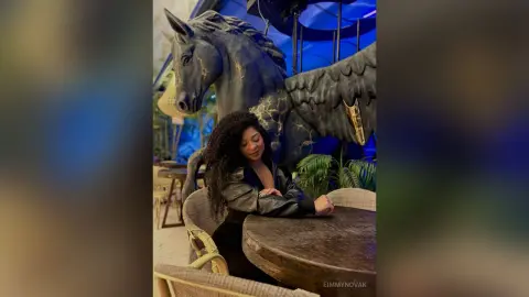Woman in a black leather jacket sitting at a cafe table near a large black Pegasus statue in a moody setting.