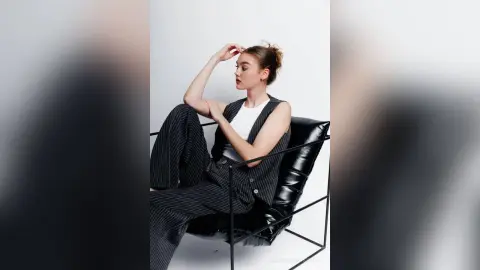 Elegant woman in a black pinstripe vest and trousers sitting on a modern black chair against a white background.
