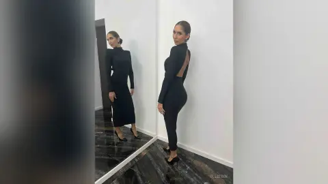 Elegant woman in a sleek black long-sleeve midi dress posing in a modern minimalist room mirror reflection.