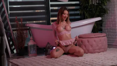 Smiling woman in pink lingerie sitting on a white rug next to a bathtub in a cozy, moody bedroom setting.