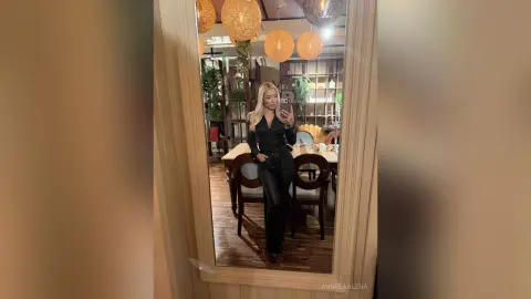 Blonde woman in a chic black outfit taking a mirror selfie in a modern, warm-lit restaurant setting.