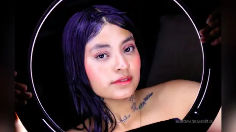 Young woman with purple hair and tattoos looking into a glowing circular mirror in a dark setting.