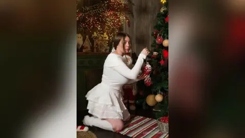 Young woman in a white festive outfit decorating a glowing Christmas tree in a cozy holiday setting.