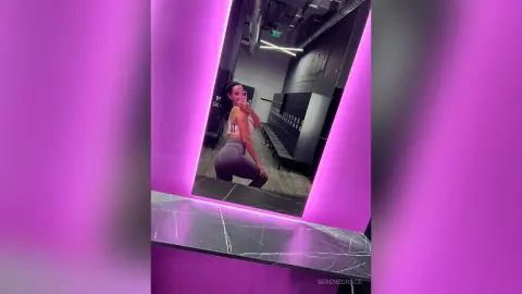 Young woman in white sports bra and grey leggings taking a selfie in a modern gym mirror with pink lighting.