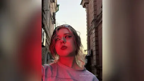 VernellZiccardi looks up with blonde hair and red lipstick while standing in an outdoor alleyway between brick buildings.