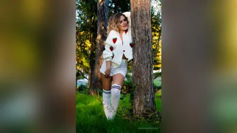 Smiling woman in white strawberry cardigan and denim shorts leaning against a tree in a sunny forest setting.
