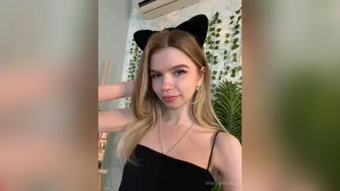 UnataMigdel wears black cat ears and a black tank top while posing in front of a leafy wall decoration.