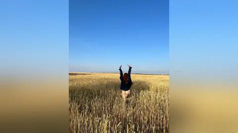 Redheaded woman in a black top raising arms in a golden wheat field under a clear blue sky.