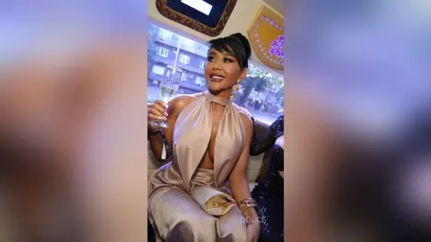 Dolce poses in a tan satin halter dress while holding a glass of champagne.
