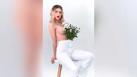 DaisyLin sits on a white stool, posing naked from the waist up with white flowers held against her chest.