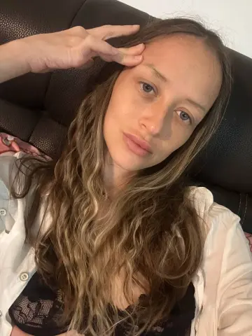 Young woman with wavy blonde hair wearing a white shirt and black lace top relaxing on a dark sofa.