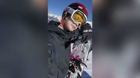 Smiling female snowboarder in a dark winter jacket and red helmet posing with her board in the sunny mountains.