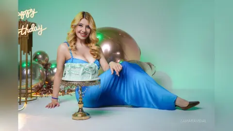 Smiling blonde woman in a blue dress sitting by a birthday cake and rose gold balloons in a festive setting.