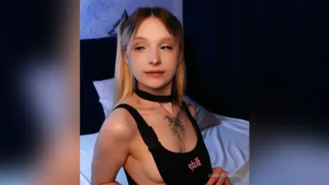 Young woman in a black tank top with chest tattoos posing in a dimly lit bedroom.