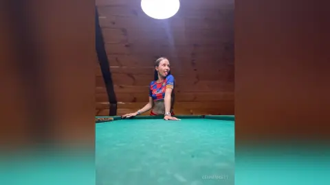 Playful young woman in a red football jersey leaning on a green pool table in a rustic wooden room.