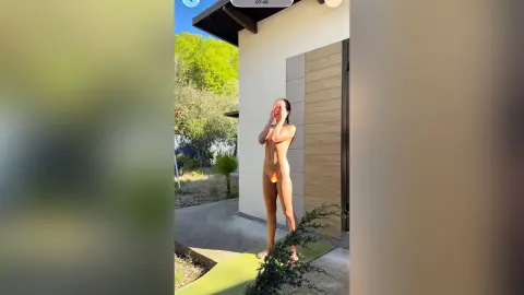 Nude woman standing outdoors by a modern house in a bright, natural garden setting.