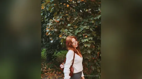 Redheaded woman in a white sweater and glasses smiling in a lush autumn forest setting.