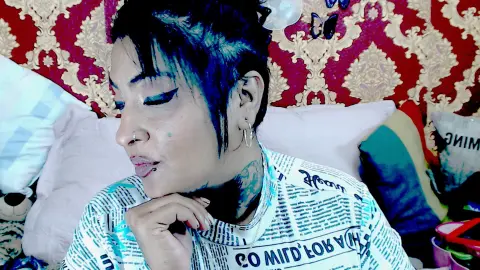 Indiancats22 looks down with a hand under her chin, showing her short black hair and nose ring while wearing a white patterned t-shirt.