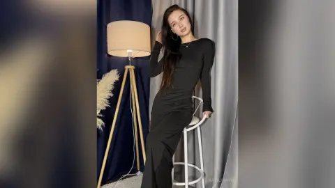 Elegant woman in a long black dress sitting on a white stool in a moody indoor studio setting.