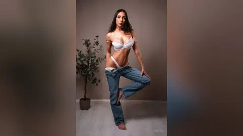 Slim brunette woman in white lingerie and blue jeans posing confidently in a minimalist indoor setting.