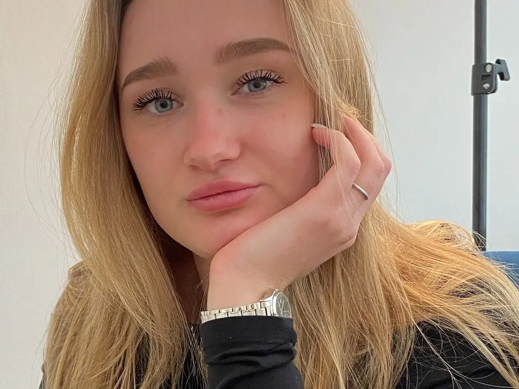 Blonde woman in a black top resting her chin on her hand with a calm expression in a bright indoor setting.