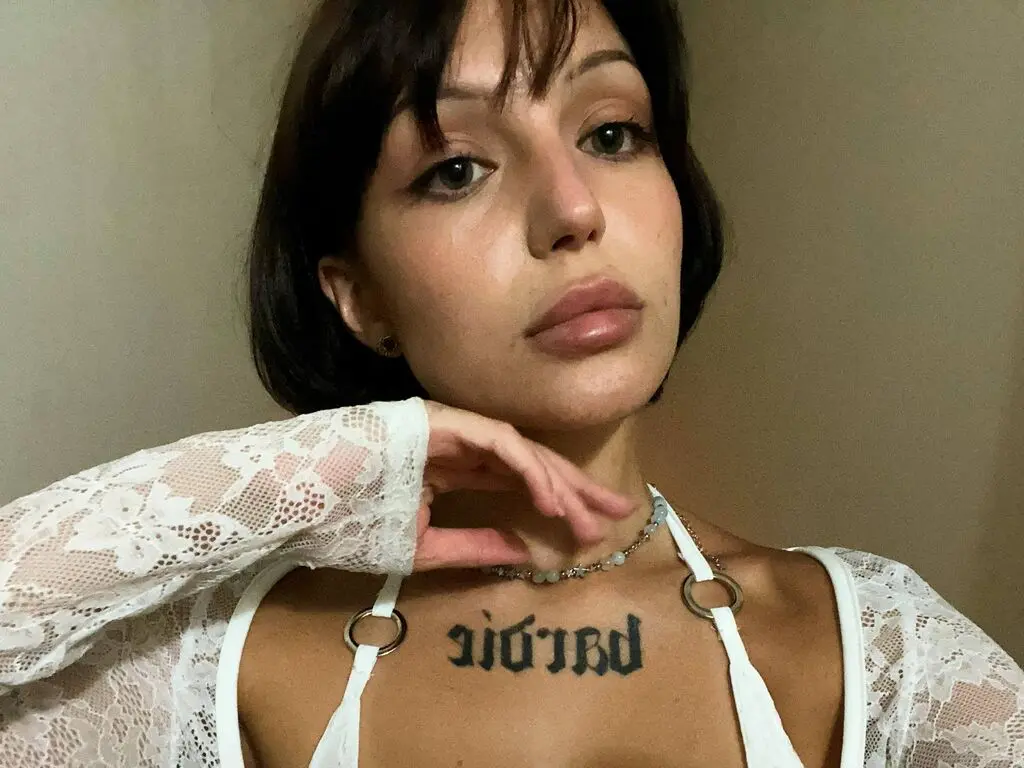 Young woman with dark hair and chest tattoo wearing a white lace top in a soft, moody indoor setting.