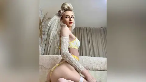Blonde woman in yellow lace lingerie posing on a white sofa in a bright, minimalist room.