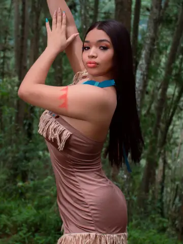 Young woman in a tan fringe dress posing gracefully in a lush green forest setting.