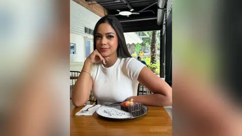 Smiling woman in a white ribbed top enjoying chocolate cake at an outdoor cafe setting.