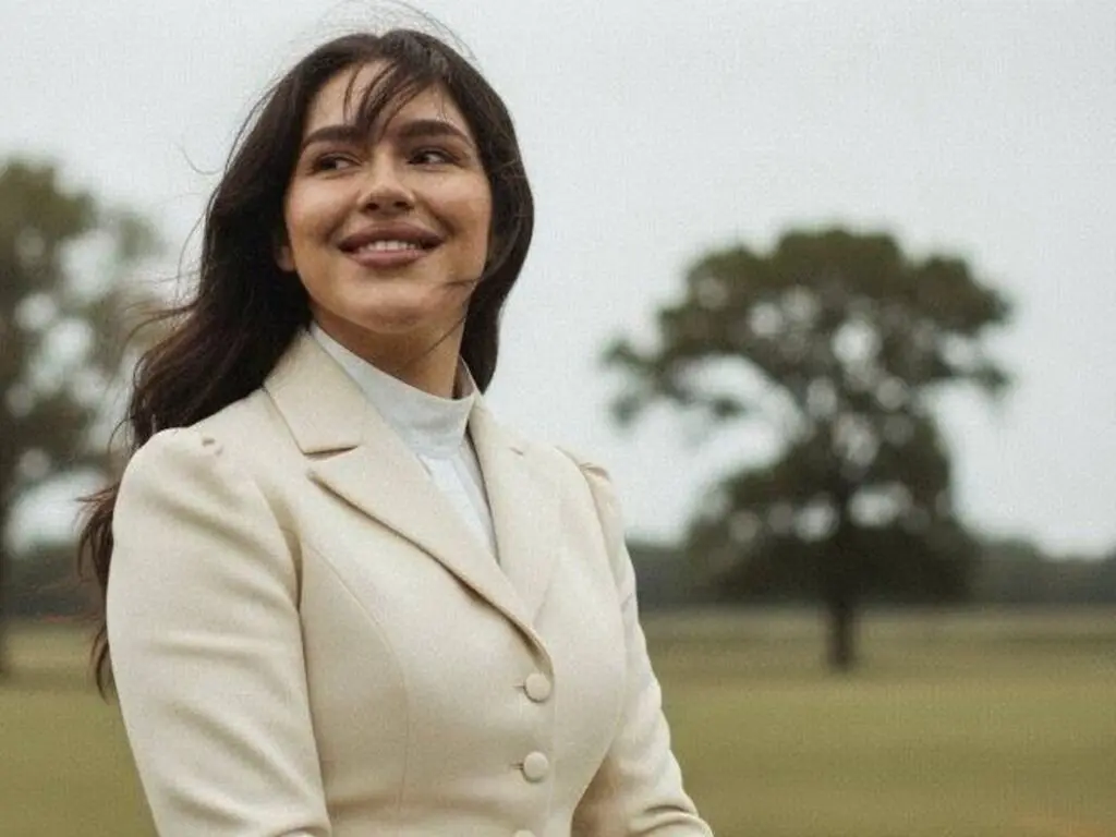 Smiling young woman in a cream blazer and white turtleneck standing in a dreamy, misty outdoor field.