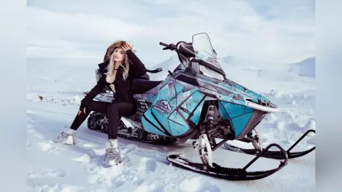 KaylaMinov poses in a black bodysuit next to a blue and white snowmobile in a snowy landscape.