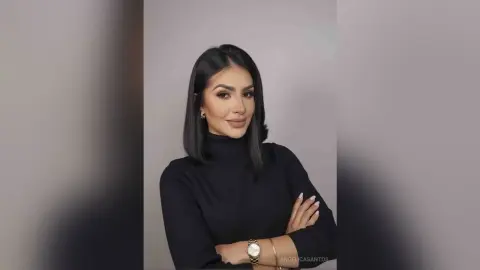 Elegant woman with dark hair in a black turtleneck sweater posing confidently against a neutral studio background.