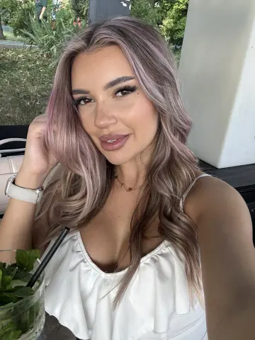 Smiling woman with wavy ash blonde hair wearing a white summer top in a bright outdoor cafe setting.