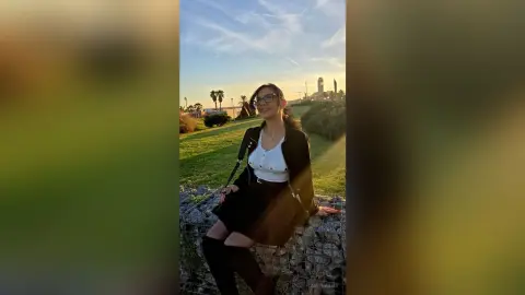 Smiling woman in a white blouse and black skirt sitting on a stone wall in a sunny park at sunset.