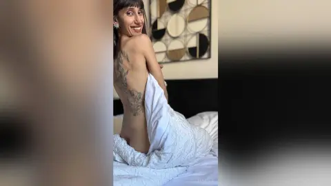 Smiling woman with back tattoos wrapped in a white textured blanket sitting on a bed in a bright room.
