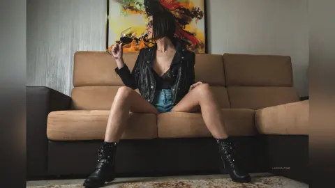 Stylish woman in a black leather jacket and denim shorts relaxing on a tan sofa with a glass of red wine.
