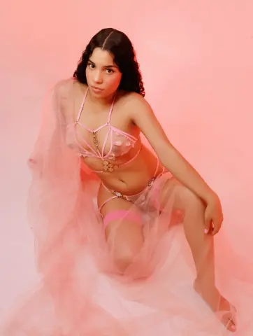 Young woman in pink lace lingerie and sheer fabric posing against a soft pink background with a dreamy mood.