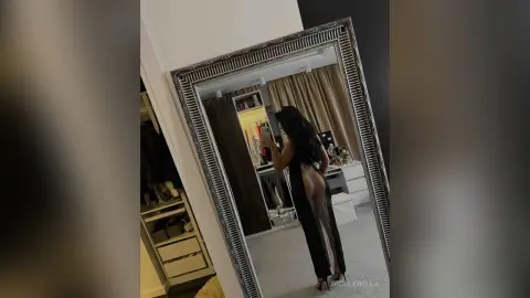 NicoleBella stands in front of a large mirror wearing a long black dress with a high slit that shows her bare leg.