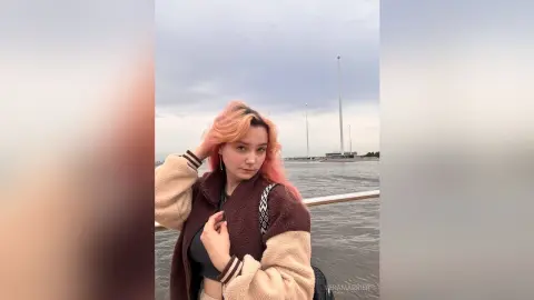 Young woman with pink hair in a cozy brown fleece jacket standing by the water under a cloudy sky.