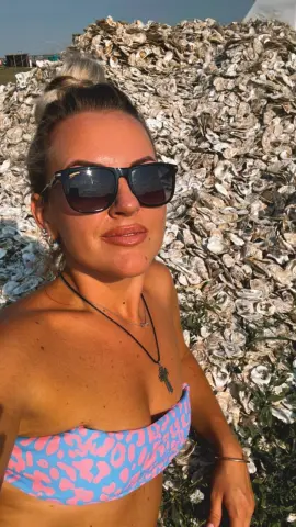 Smiling woman in pink leopard print bikini and sunglasses posing in front of a large pile of oyster shells.