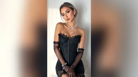Elegant woman in a black lace corset dress and pearl necklace posing in a bright, minimalist indoor setting.
