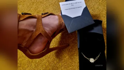 Elegant brown suede strappy high heel sandals next to a black gift box and jewelry on a textured gold surface.