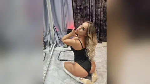 Blonde woman in a black bodysuit sitting on a white chair in a modern room with a moody atmosphere.